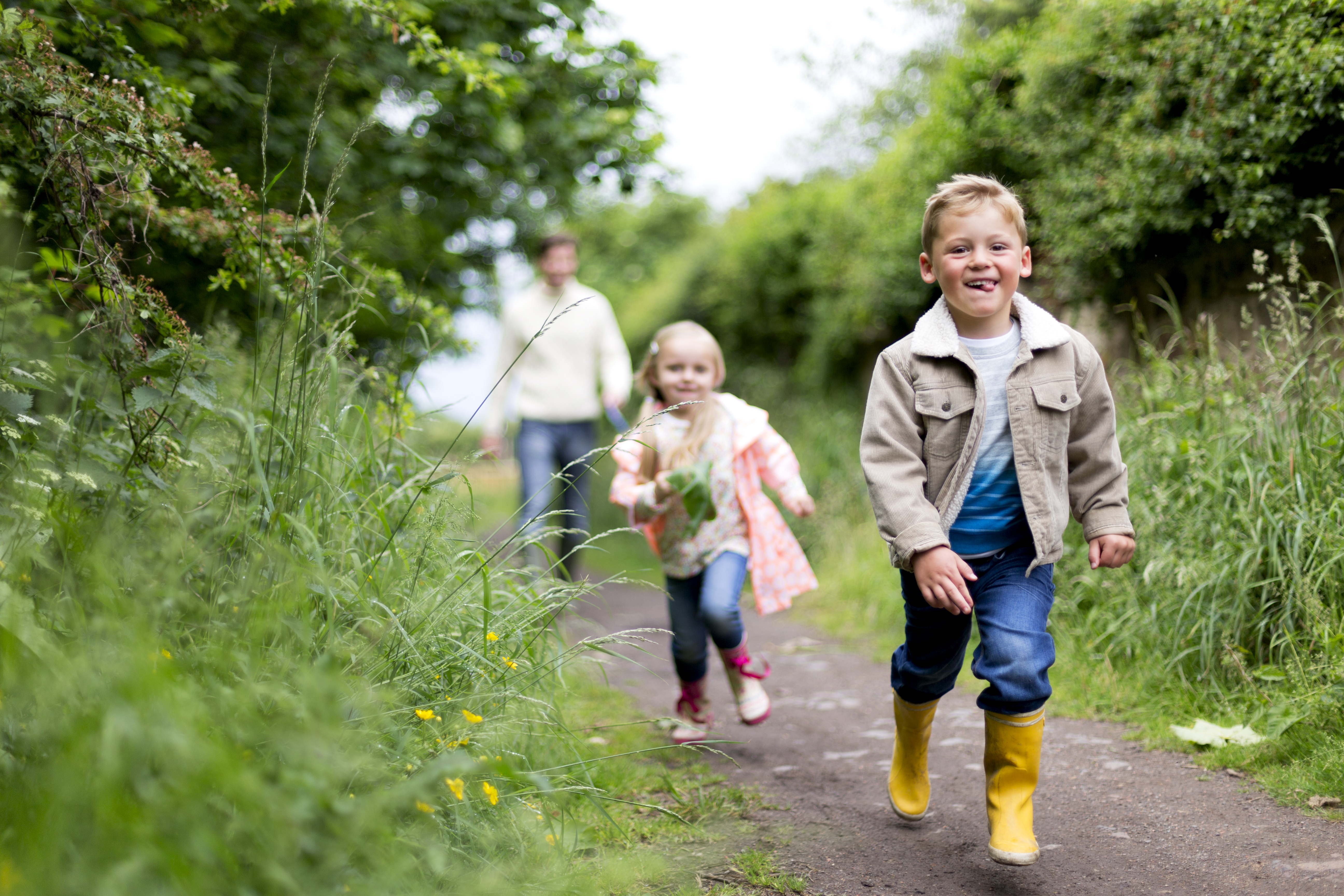 Children running down a country dirt track ahead of their father. Happily enjoying time together. Boy runs ahead of them all as the little girl take chase.