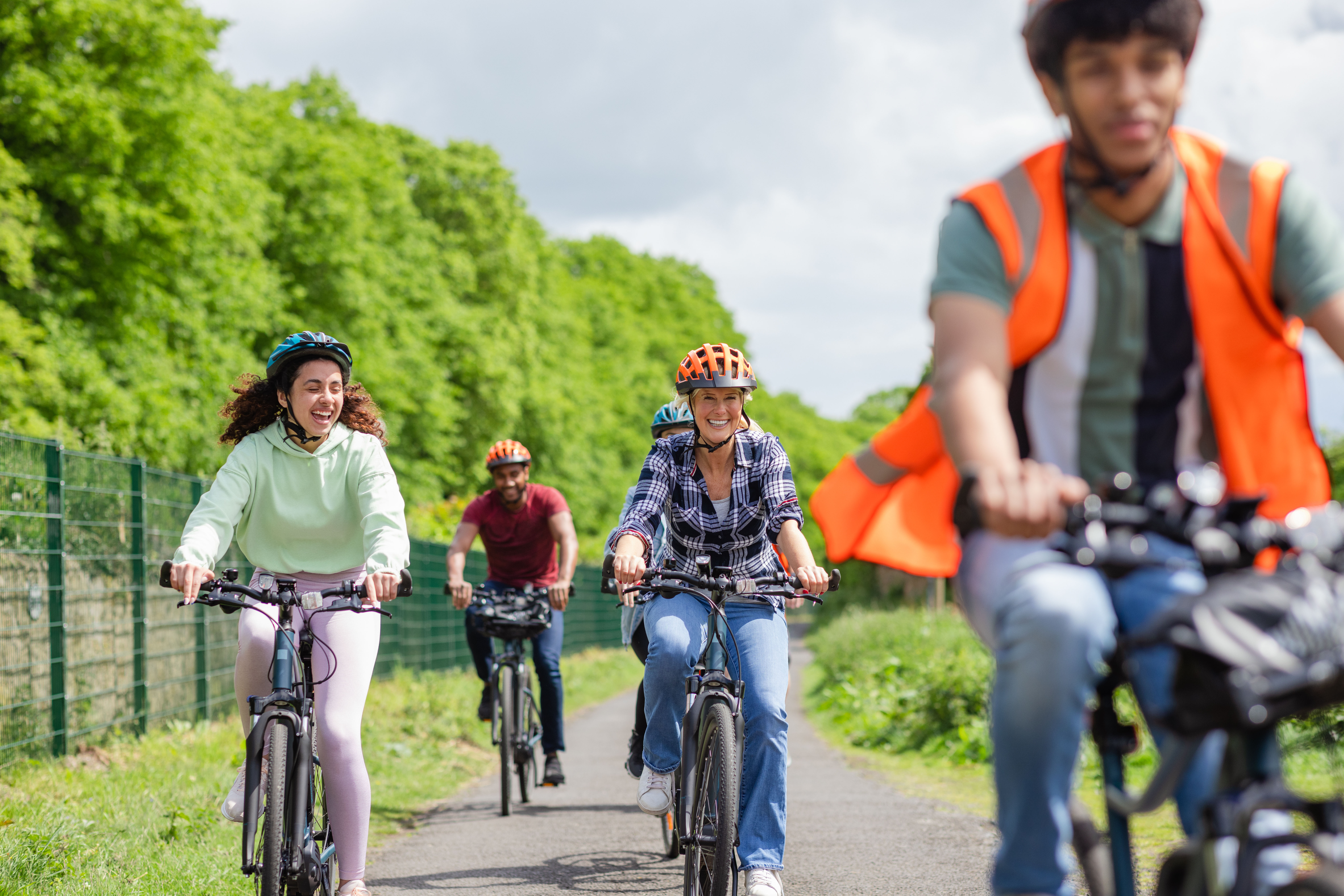 A head-on shot of a small group of people cycling along a path whilst on a community bicycle ride in Hexham, North East England. They are smiling as they cycle, wearing casual clothing and bicycle helmets. The male at the front is wearing a reflective waistcoat.